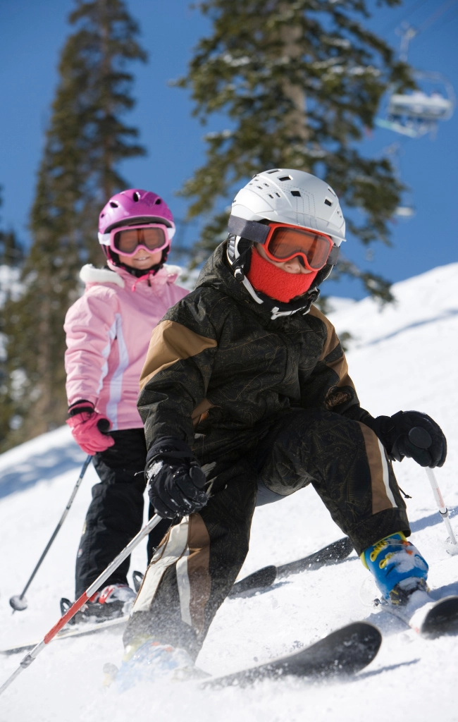 Two children skiing downhill wearing helmets and winter gear on a snowy mountain